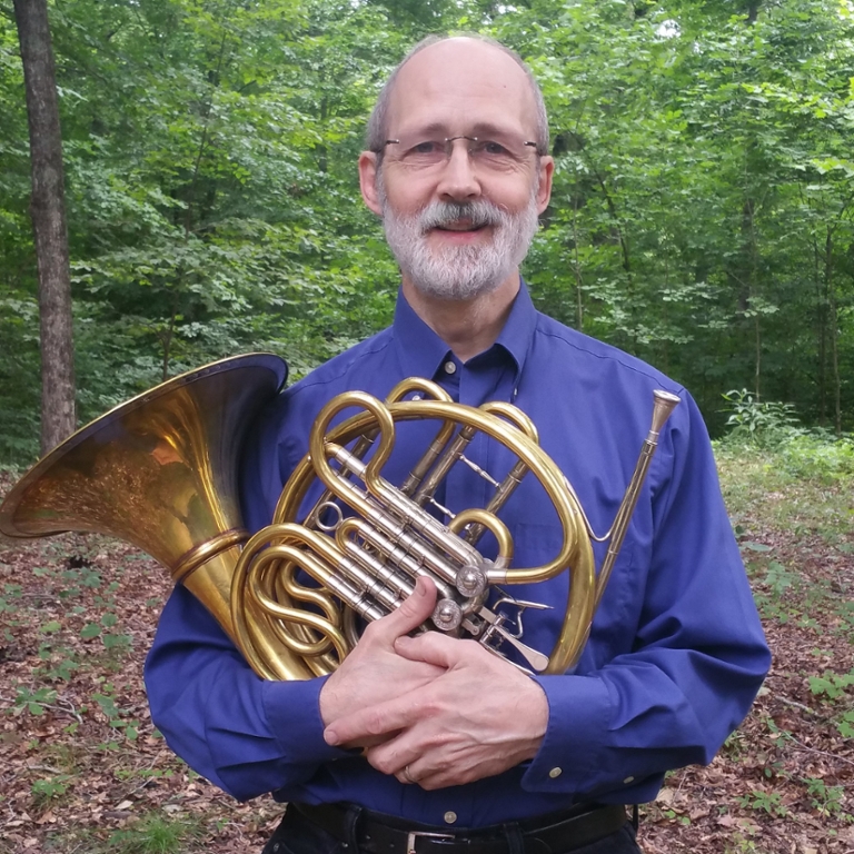 a bearded man holding a french horn in the woods