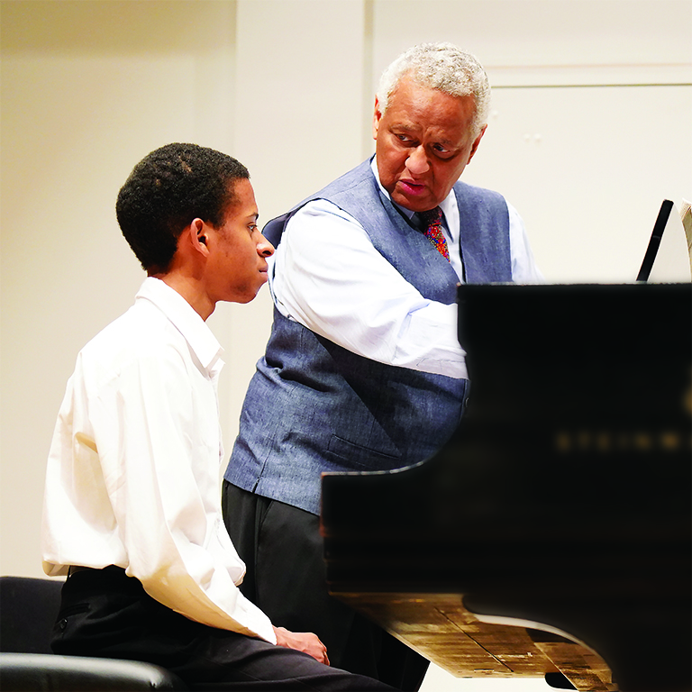 Student in a white shirt sitting at a piano while a faculty member provides instruction.