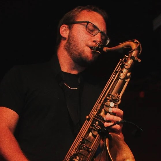 Man wearing a black shirt and glasses, playing a saxophone. His eyes are closed.