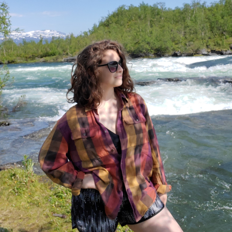 Woman wearing sunglasses, a red and black flannel, and shorts, standing outside near a river with trees and a mountain in the background.