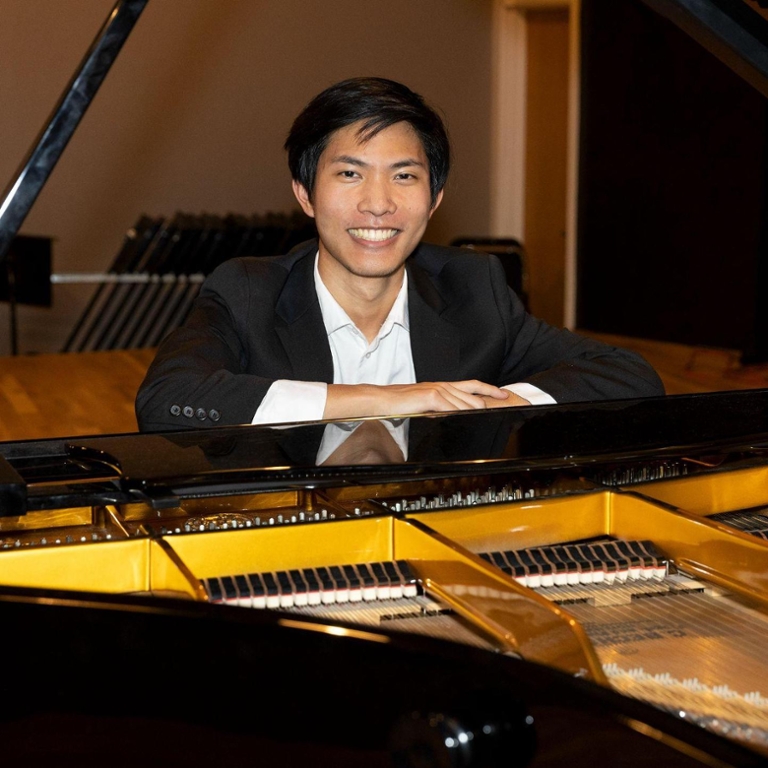 Hans-Derek Yu, sitting at a piano smiling and wearing a tuxedo
